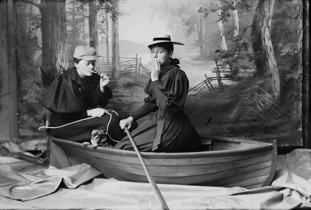 Two women sitting in a row boat in a photography studio. One smokes.