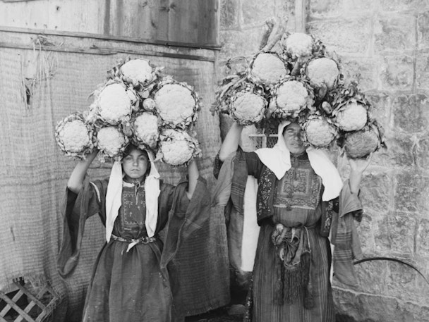 Two women in traditional dress carry large bundles of cauliflower on their heads while standing against a stone wall.