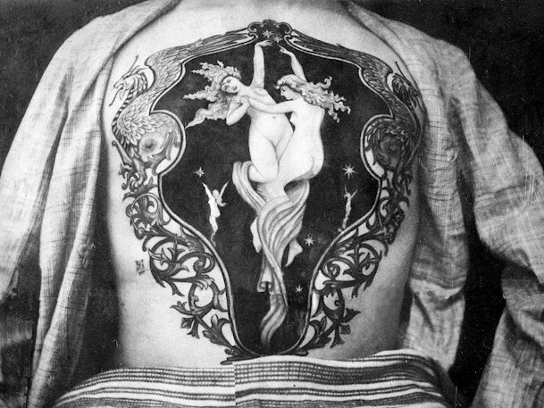 Exposed back shows large tattoo depicting two nude figures dancing among stars, surrounded by an ornate frame with flowing scrollwork designs.