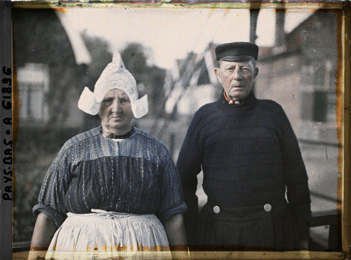 Dutch couple in traditional dress - woman wearing white cap and striped dress with apron, man in dark uniform with captain's hat standing together.