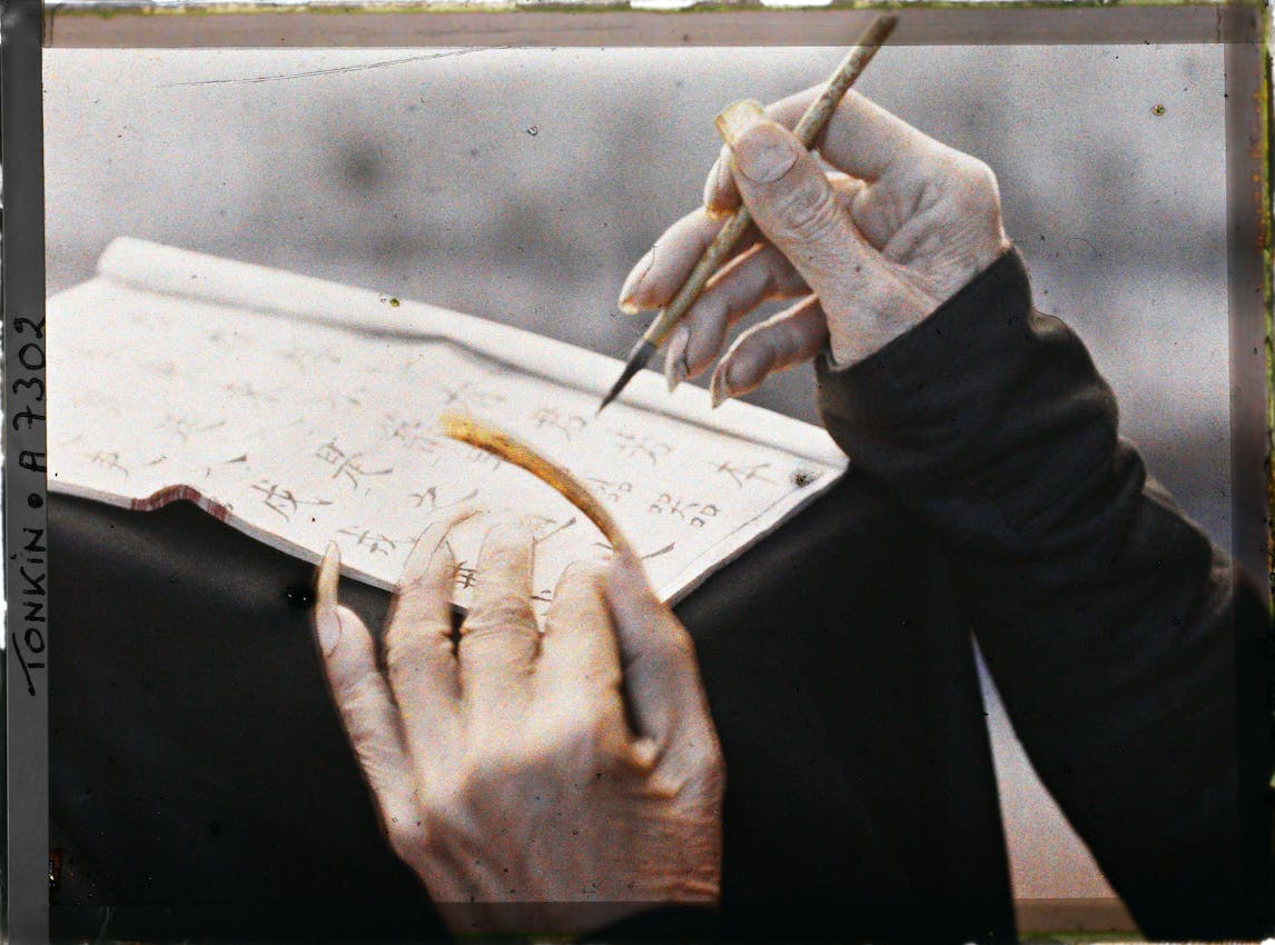 Detailed view of calligraphy in progress showing two hands at work - right hand carefully controlling a brush over paper, left hand stabilizing the document, both hands emerging from dark sleeves.