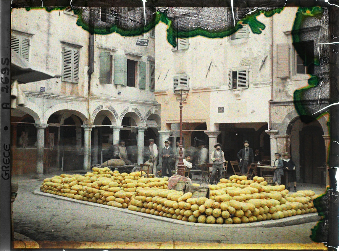 Men gather under arched colonnade of Mediterranean building with large display of stacked yellow melons in courtyard, street lamp mounted on wall.