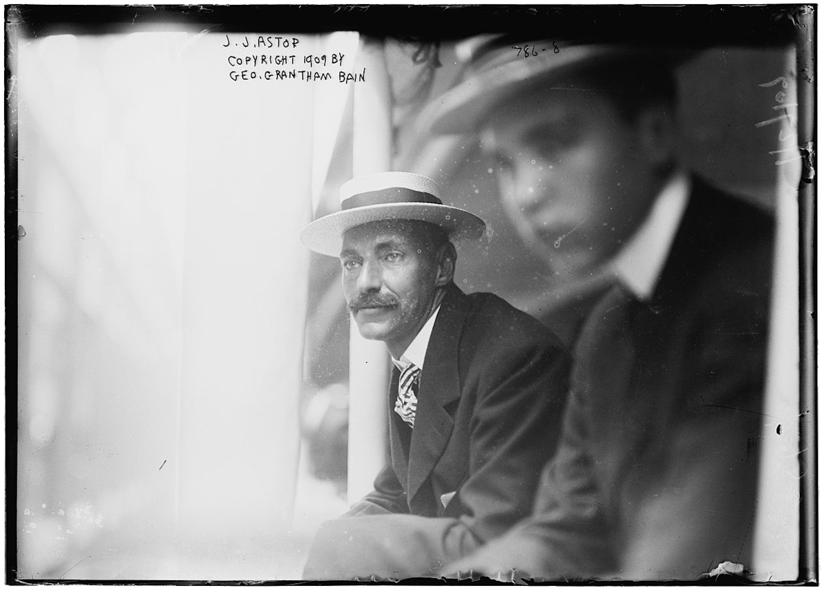Astor leans from a train window in the center of the image wearing a straw boater hat, a suit with tie, and a mustache