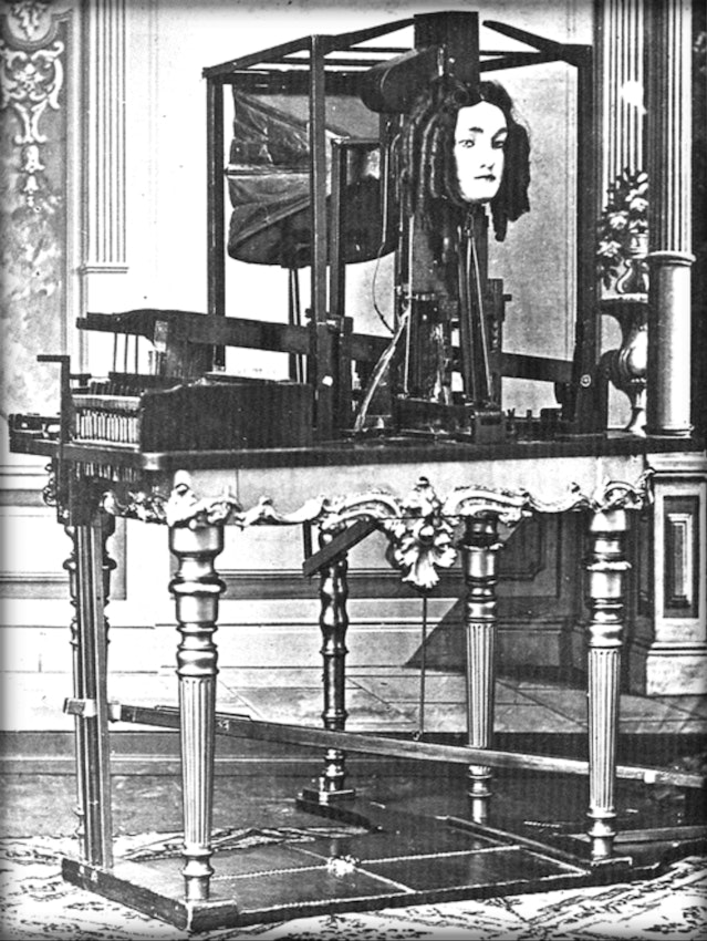 A mechanical device featuring a realistic human head with moving parts, mounted on an ornate table with various mechanisms visible.
