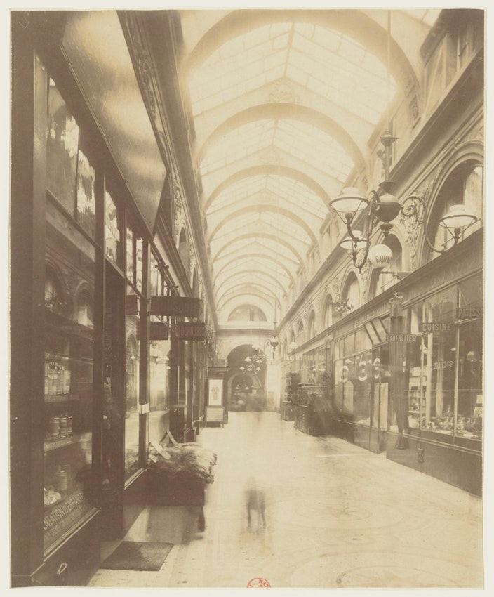 Sepia-toned photograph of an elegant indoor shopping arcade with a glass-vaulted ceiling, gas lamps, and detailed shop fronts. A blurred figure, possibly due to long exposure, adds a ghostly presence to the scene.