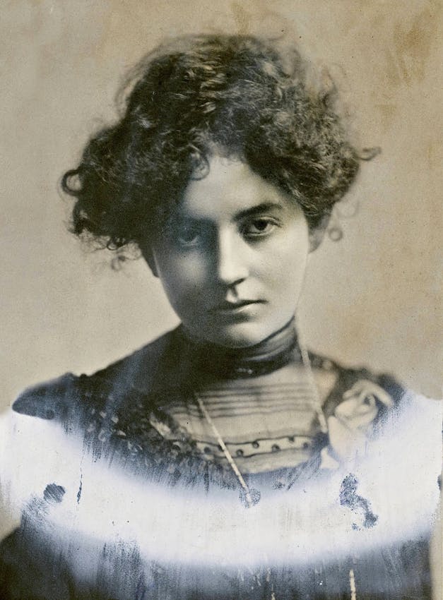 Close-up portrait of a woman with tousled dark curly hair wearing a high-necked blouse or dress with detailed collar and subtle embellishments. The photograph has visible aging marks and minor deterioration around the edges.