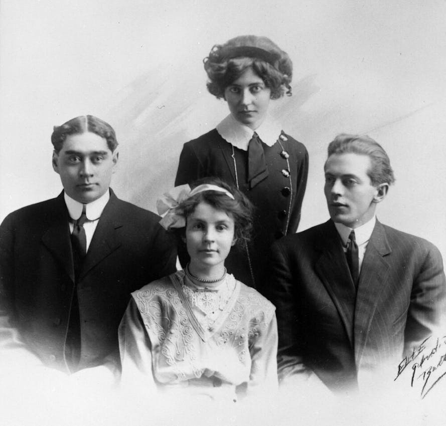 Formal studio portrait of four young people arranged in a composition with two men in dark suits with high collars flanking two women - one seated in front wearing an ornate white blouse with embroidery and a hair accessory, another standing behind in a dark dress with white collar.