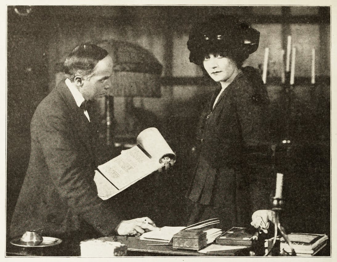 A man in a suit examines an open book while standing at a desk with Mary MacLane, who wears a dark dress and hat, looking directly at the camera while standing beside him.