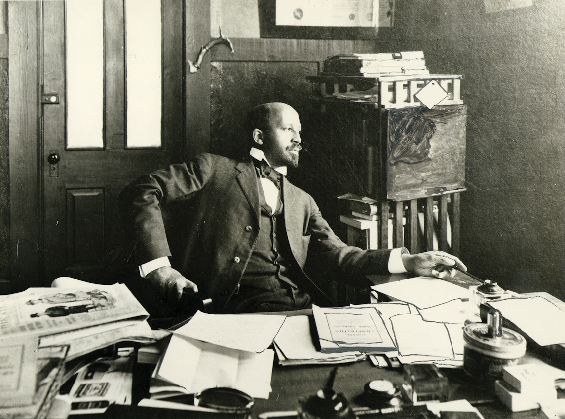 Man with goatee in suit sitting at desk covered with papers and documents, looking to the side with papers and filing system visible behind him.