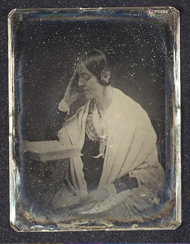 A person in a light-colored garment sits in profile view, head resting thoughtfully on one hand, with dark hair styled back