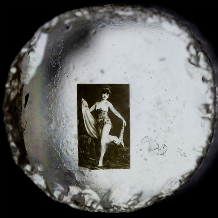 Small circular view around a rectangular photograph of a woman in performance costume holding fabric while posing with one leg raised against a dark background."