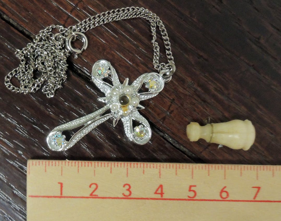 Silver cross pendant necklace with pearl center and crystal accents on a chain, displayed next to a small ivory chess piece and ruler showing size.
