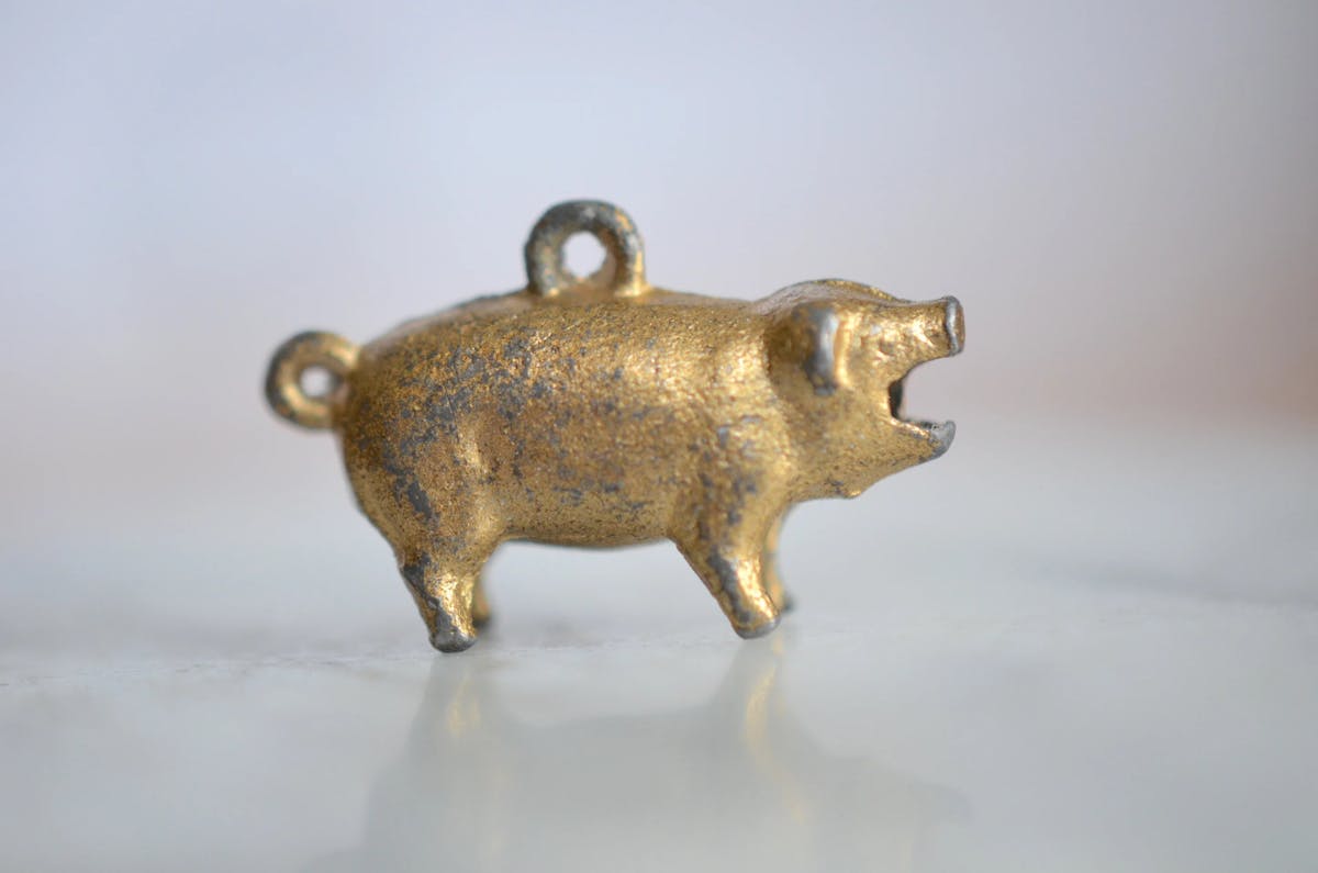 Small brass or bronze pig pendant with two hanging loops on its back, mouth open as if squealing, displayed against a light gray background.