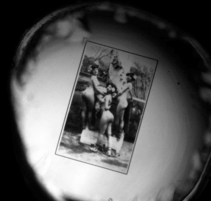 Black and white circular view through optical device showing an antique photograph of nude figures in a garden or outdoor setting with foliage visible in the background.