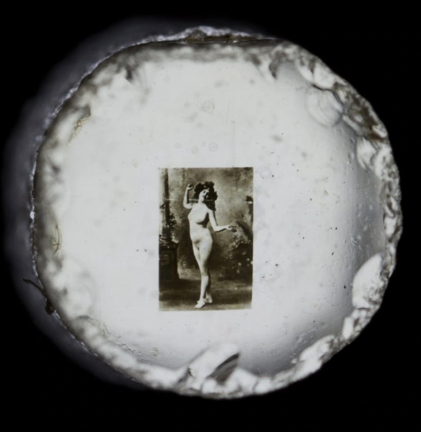 Circular view through an optical device showing a nude figure study photograph mounted on a white surface with rough edges visible around the circular frame.