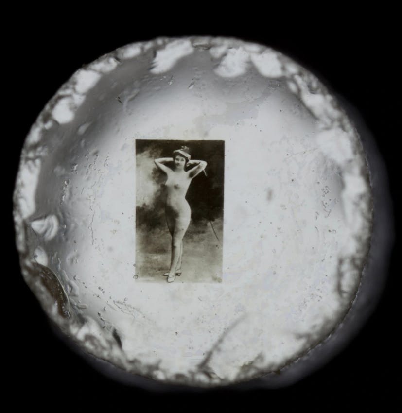 Circular view through an optical device showing a vintage figure study photograph mounted on a white surface with rough edges visible around the circular frame.