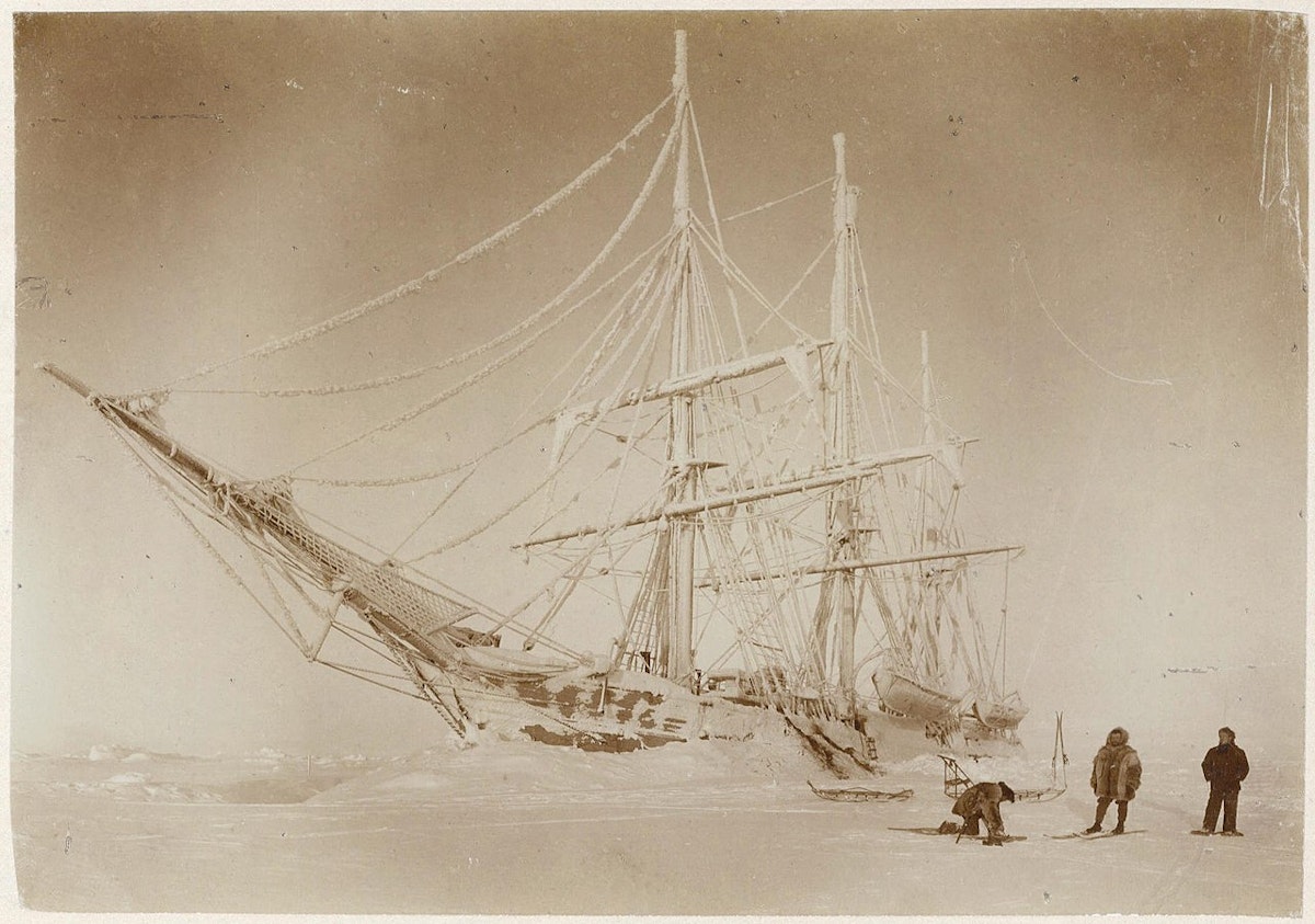 black and whote photo of a ship stuck in the ice, three explorers wearing snowshoes and skis to the right