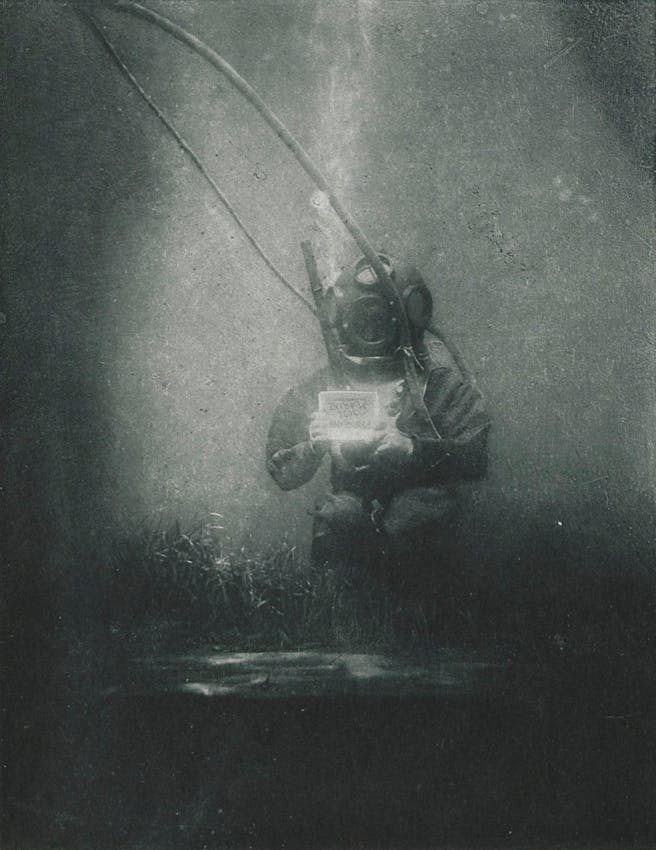 man in diving suit on the sea bed holding a written sign in his hand, the writing hard to make out