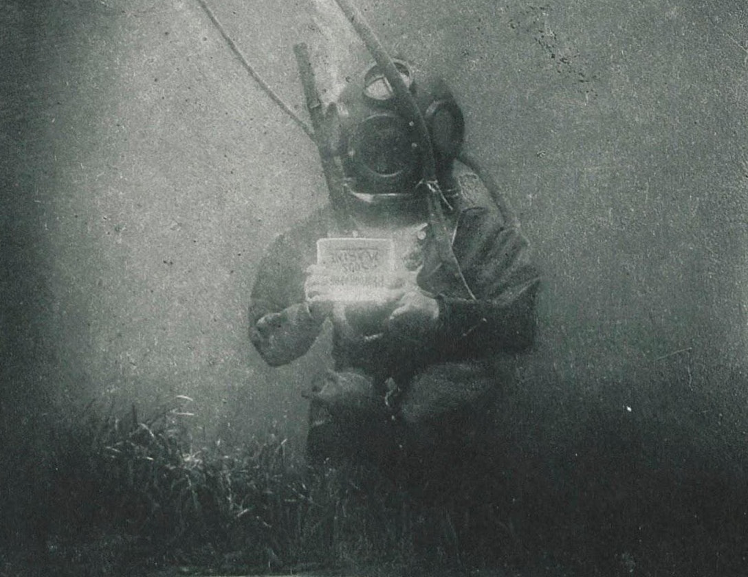 man in diving suit on the sea bed holding a written sign in his hand, the writing hard to make out