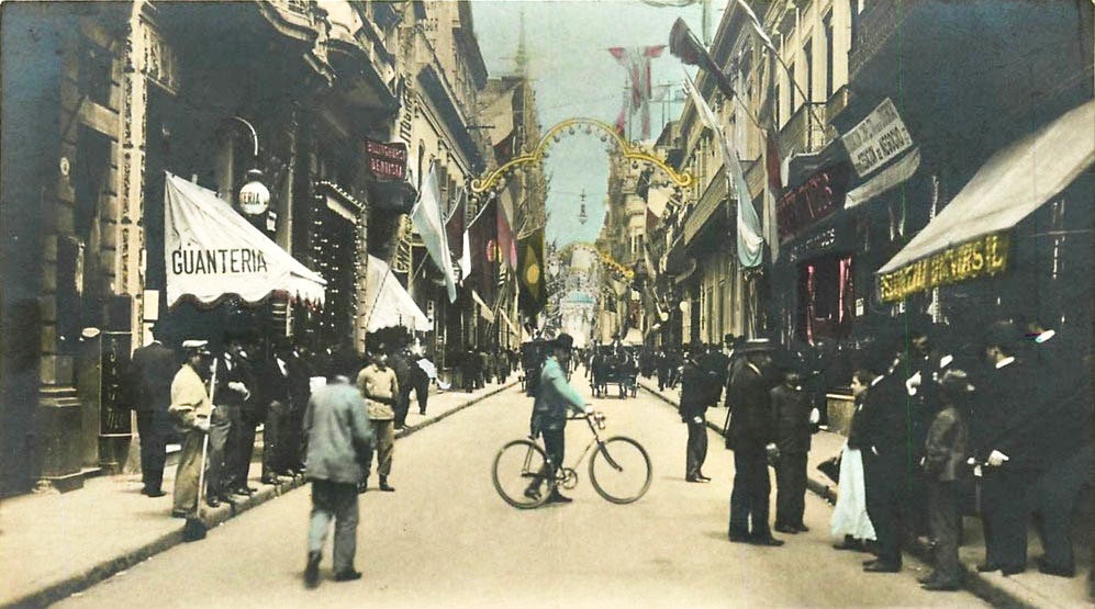 A street scene with a man on bicycle looking at the camera and crowds in front of busy shops