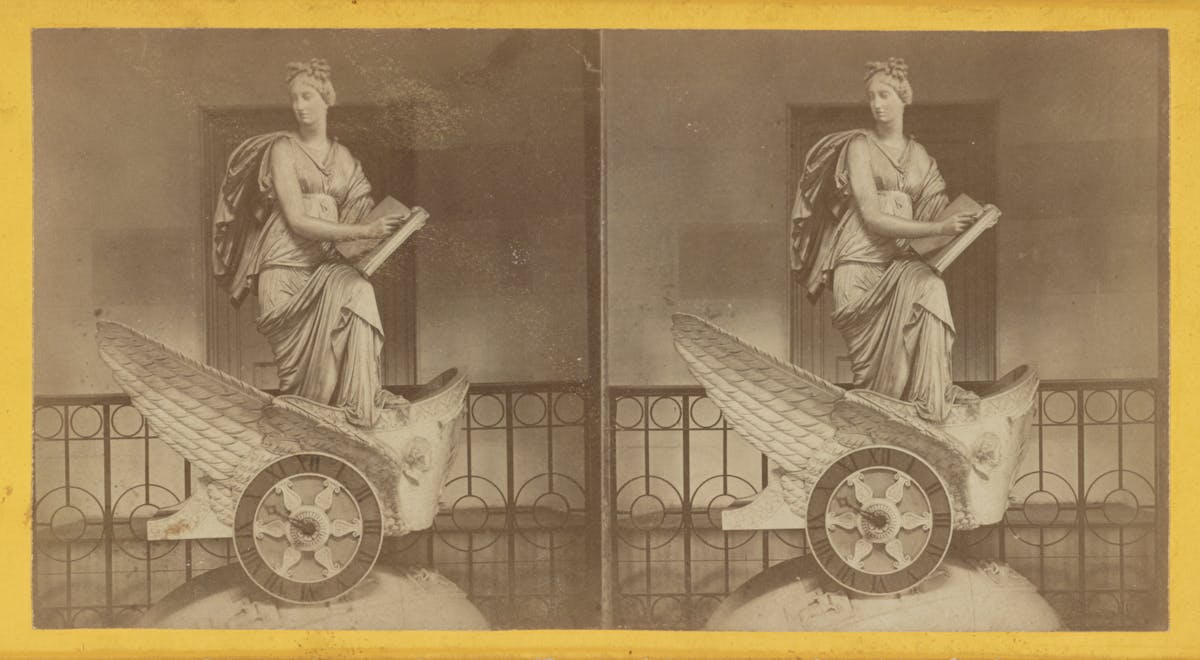 The statue depicts a woman in classical attire, seated on a winged chariot with intricate details, including feathered wings and a patterned wheel. She is holding a tablet and stylus, appearing thoughtful and serene. The statue is positioned in an indoor setting with a decorative railing and an open doorway in the background.