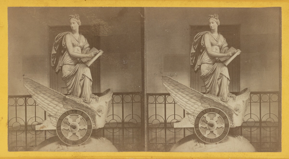 The statue depicts a woman in classical attire, seated on a winged chariot with intricate details, including feathered wings and a patterned wheel. She is holding a tablet and stylus, appearing thoughtful and serene. The statue is positioned in an indoor setting with a decorative railing and an open doorway in the background.