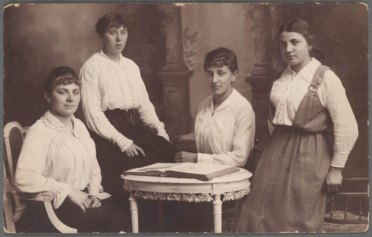 Four women in white blouses pose around a small table with an open book.