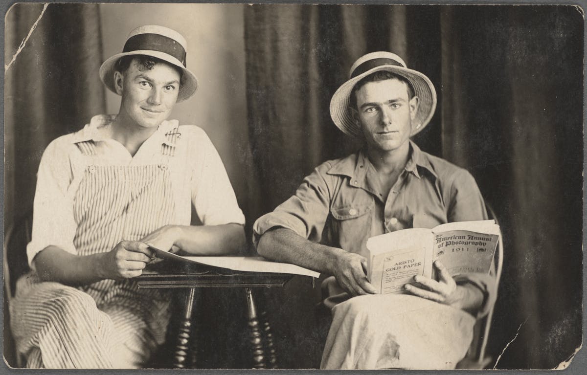 Two men wearing straw hats sit at a table, one reading a magazine and the other smiling at the camera.