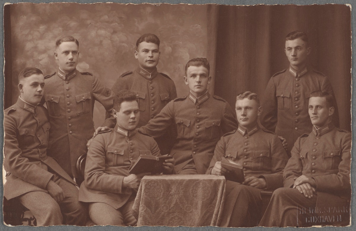 Eight men in military uniforms pose for a group portrait, some seated with books and others standing behind them.