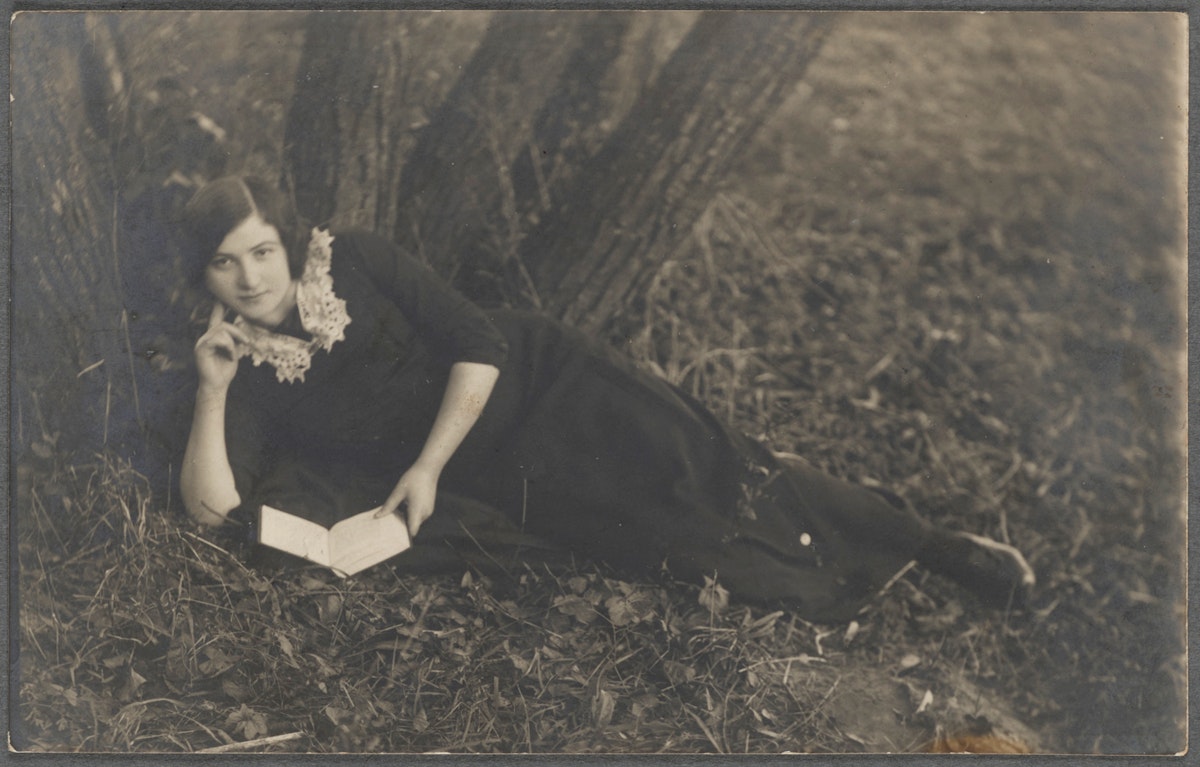 A woman in a dark dress with a lace collar reclines on the ground outdoors, holding an open book.