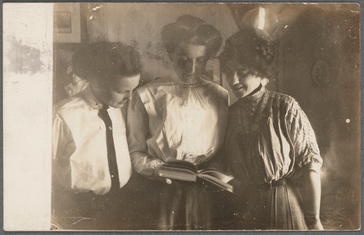 Three women standing close together, looking at an open book, smiling.