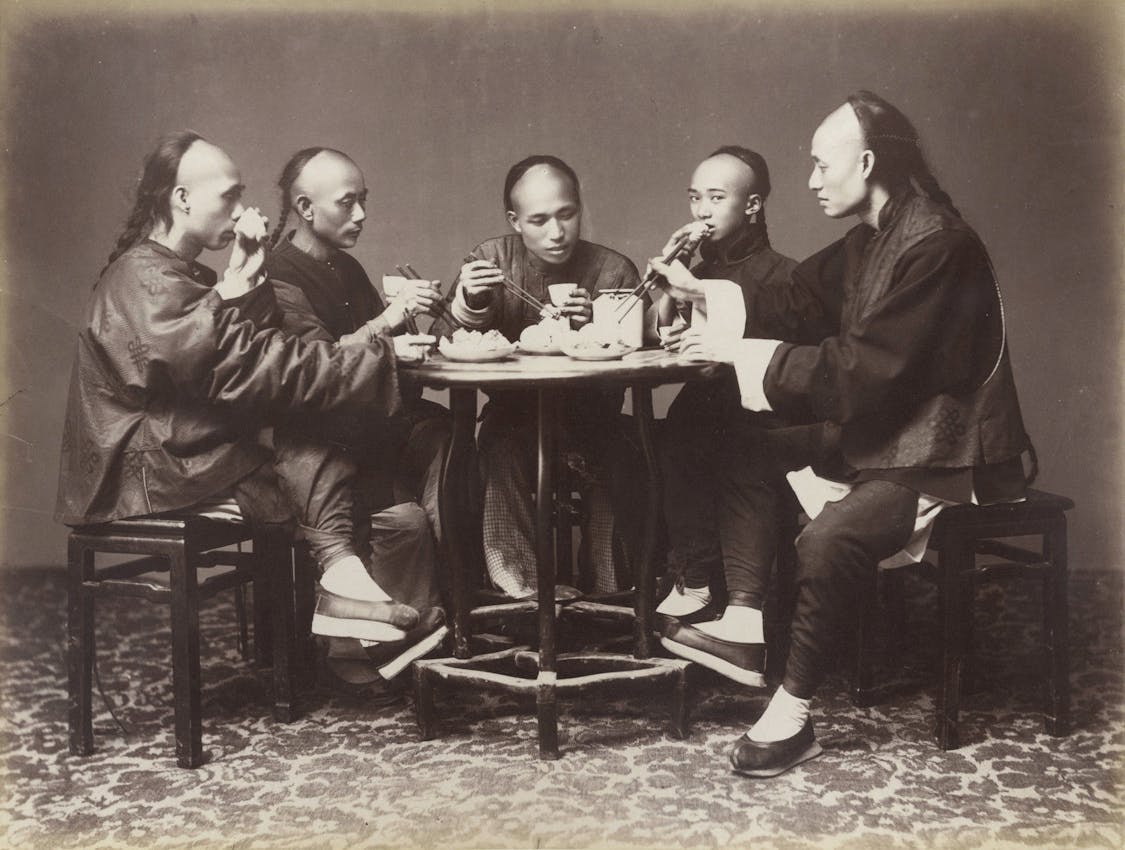 Five men wearing traditional Chinese clothing with queue hairstyles gathered around a circular table for a meal, with teacups, bowls, and chopsticks visible on the table surface, seated on wooden chairs against a plain backdrop