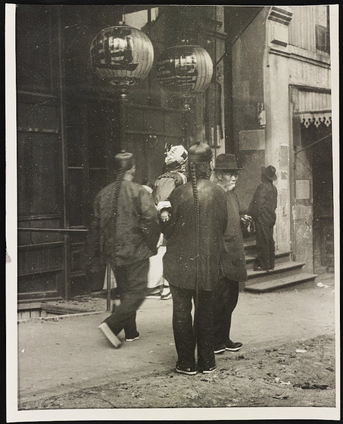 Black and white street scene showing several people in dark clothing walking along a sidewalk past storefronts decorated with round Chinese lanterns