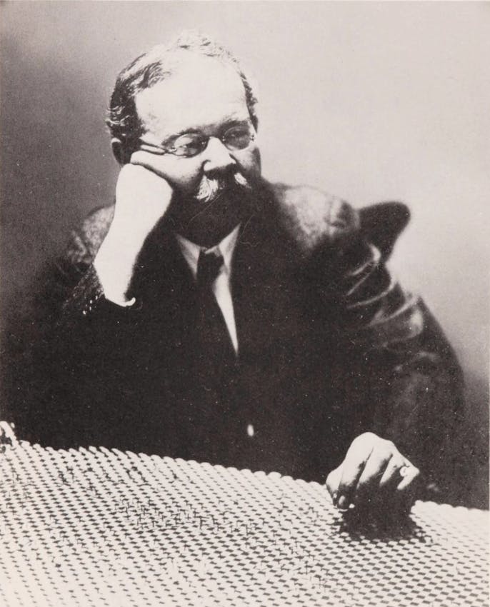 A mustachioed man in glasses rests his head on his hand while examining a textured surface covered in small, uniform objects and markings.