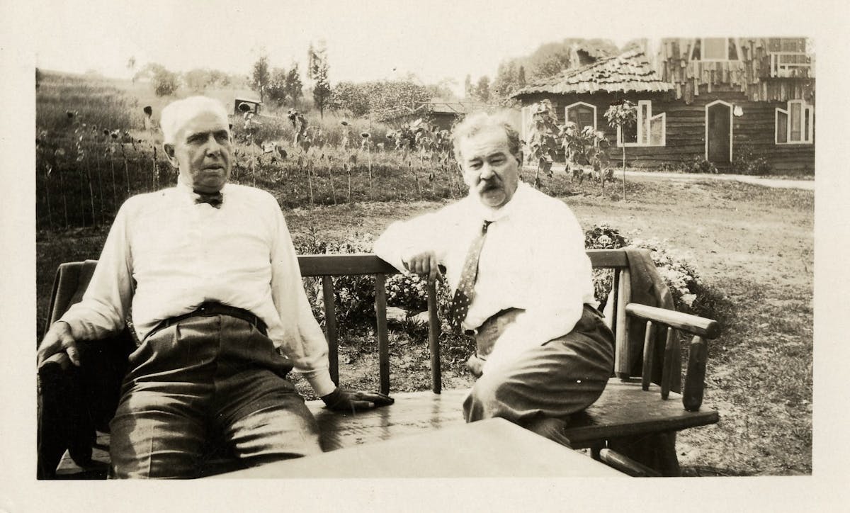 Two older men in white shirts sit on wooden chairs outdoors, with a rustic cabin and flower garden visible in the background.