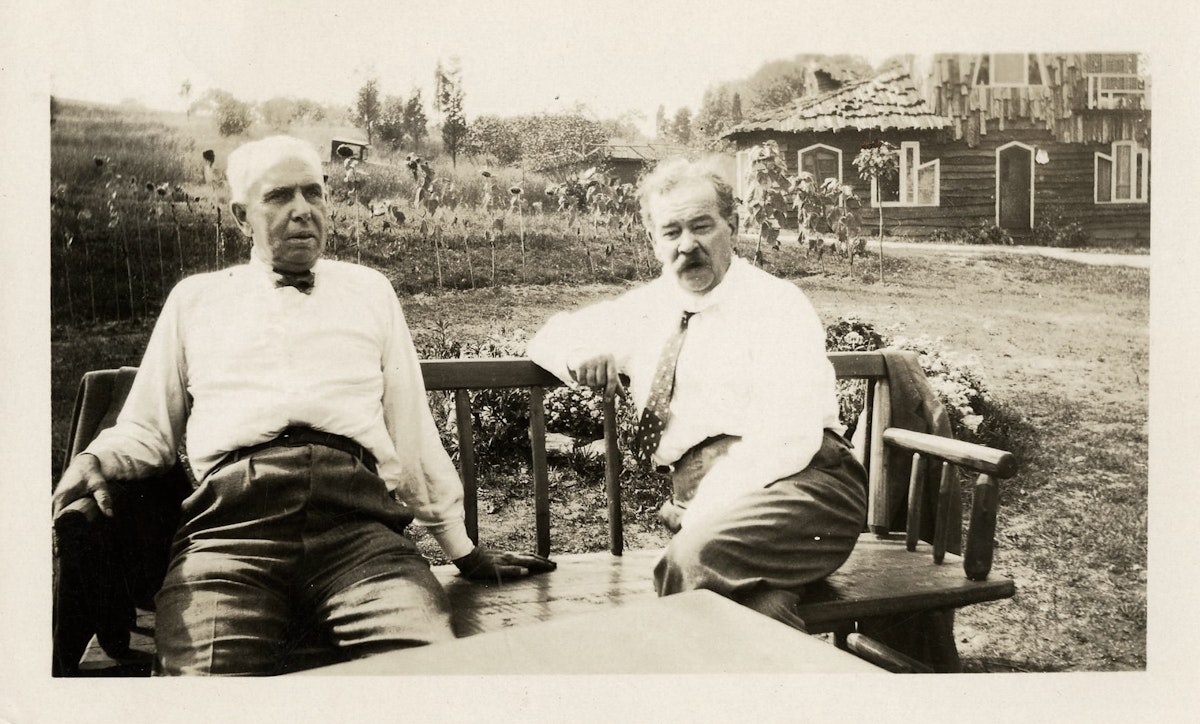 Two older men in white shirts sit on wooden chairs outdoors, with a rustic cabin and flower garden visible in the background.