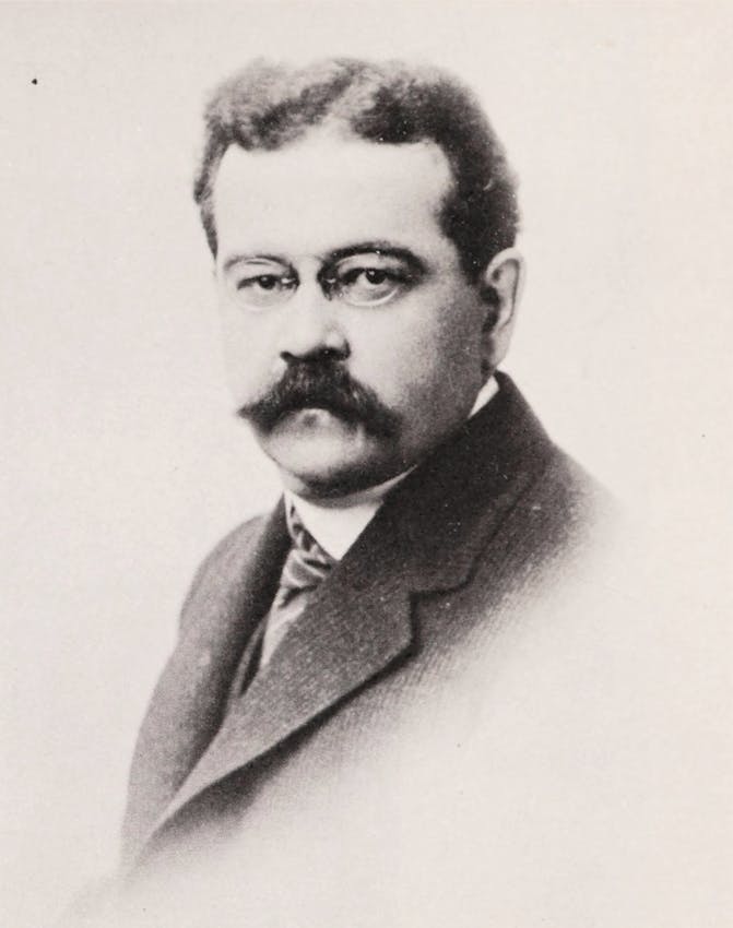 Head and shoulders portrait of a gentleman with a prominent mustache and intense gaze, wearing a dark suit with striped tie and prominent lapels against a plain background.