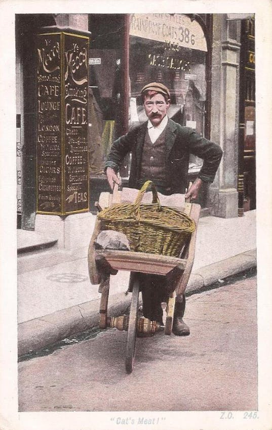 A street vendor with a flat cap and mustache poses with his wheelbarrow outside a cafe lounge advertising coffee and teas on decorative pillars.