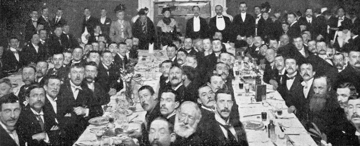 A large group of men in formal attire with bow ties and mustaches seated at long tables for a banquet dinner with place settings and glassware.