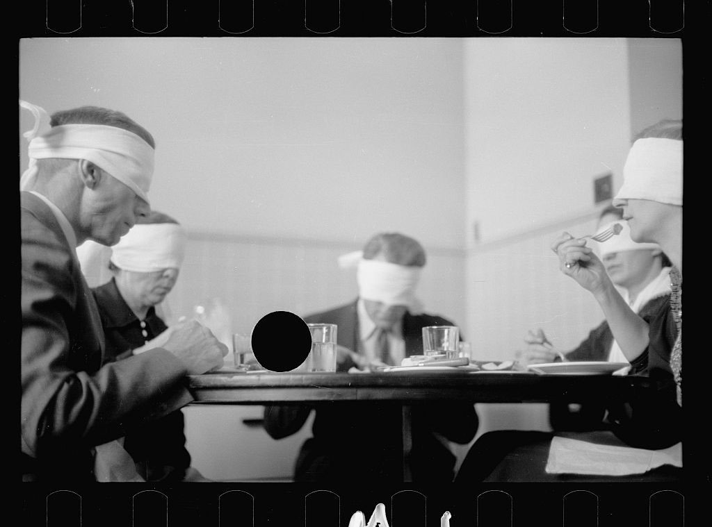 black and white photo of a table with blindfolded people eating, putting forks to their mouths, a punctured hole through the glasses on the table