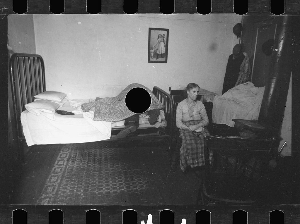 sad woman sits by a bed, on which a man lies under a blanket, only his legs visible, and the punctured hole through his body 