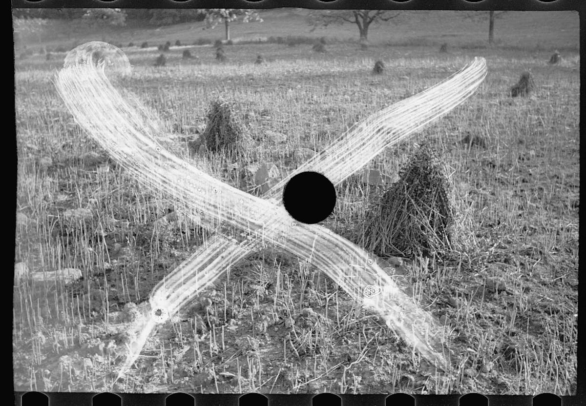 Black and white photograph of a crop field, triangular grain stacks dotted about, with a large white cross and punctured hole overlaid