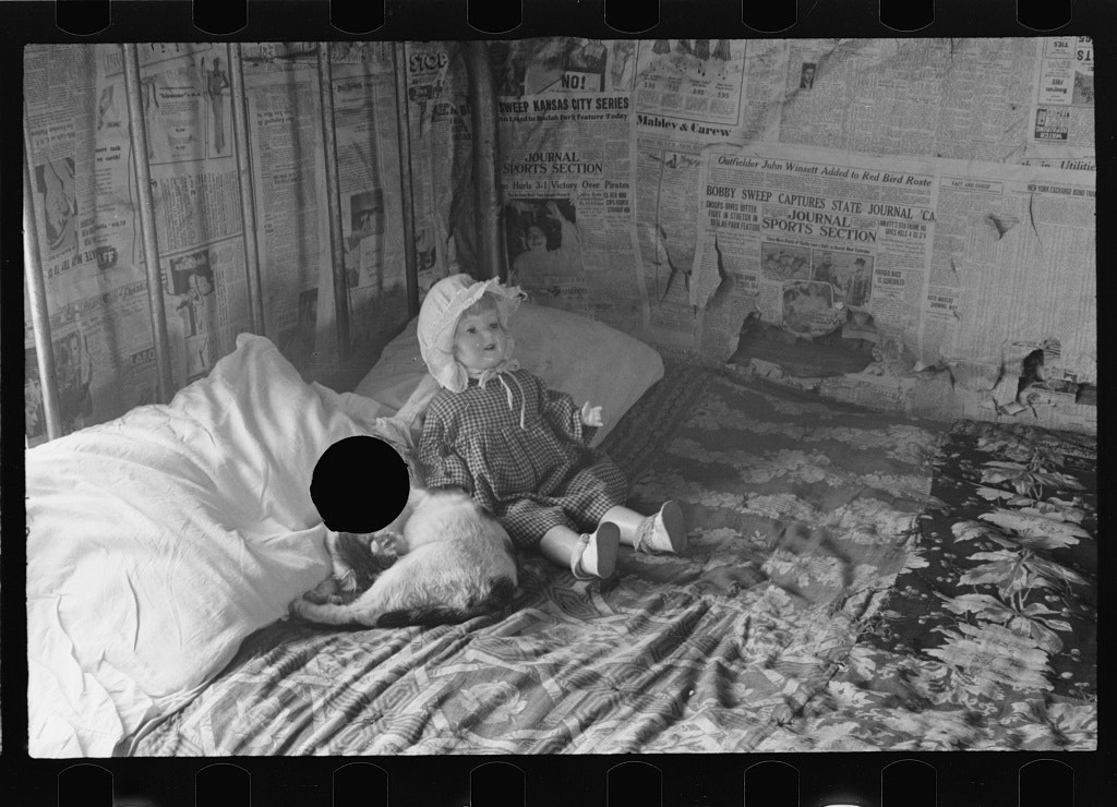black and white photo of a bedroom, was papered with newspaper, on the bed a doll and a cat, the punctured hole through the cat