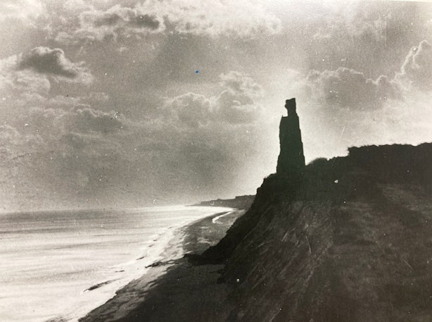 Black and white photo of a rugged coastline with a distinctive rock formation resembling a tower, under a dramatic sky with visible clouds, next to a calm sea.