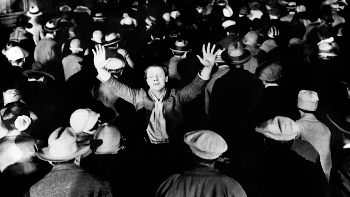 A person stands in a crowd with arms raised, facing forward while the people around them have their backs turned.