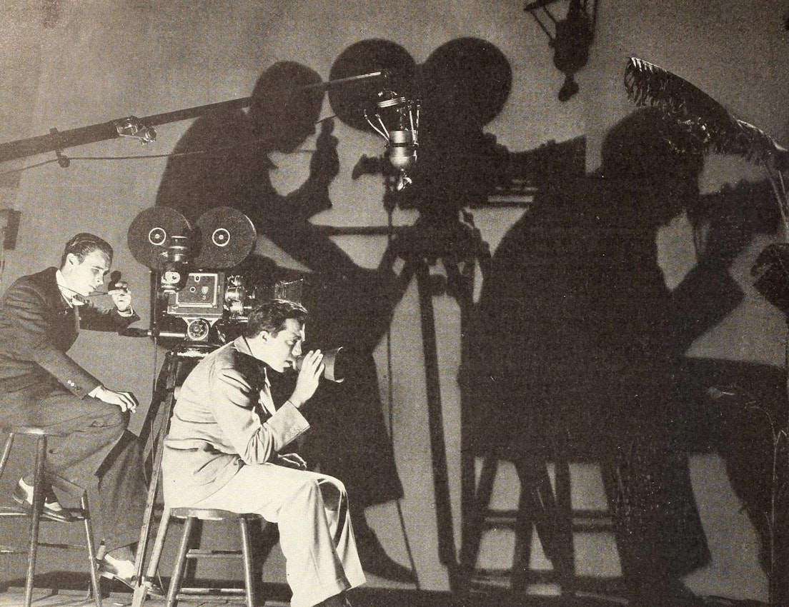 Two men work with a large film camera on set, their shadows dramatically cast on the wall behind them as they focus on the equipment.