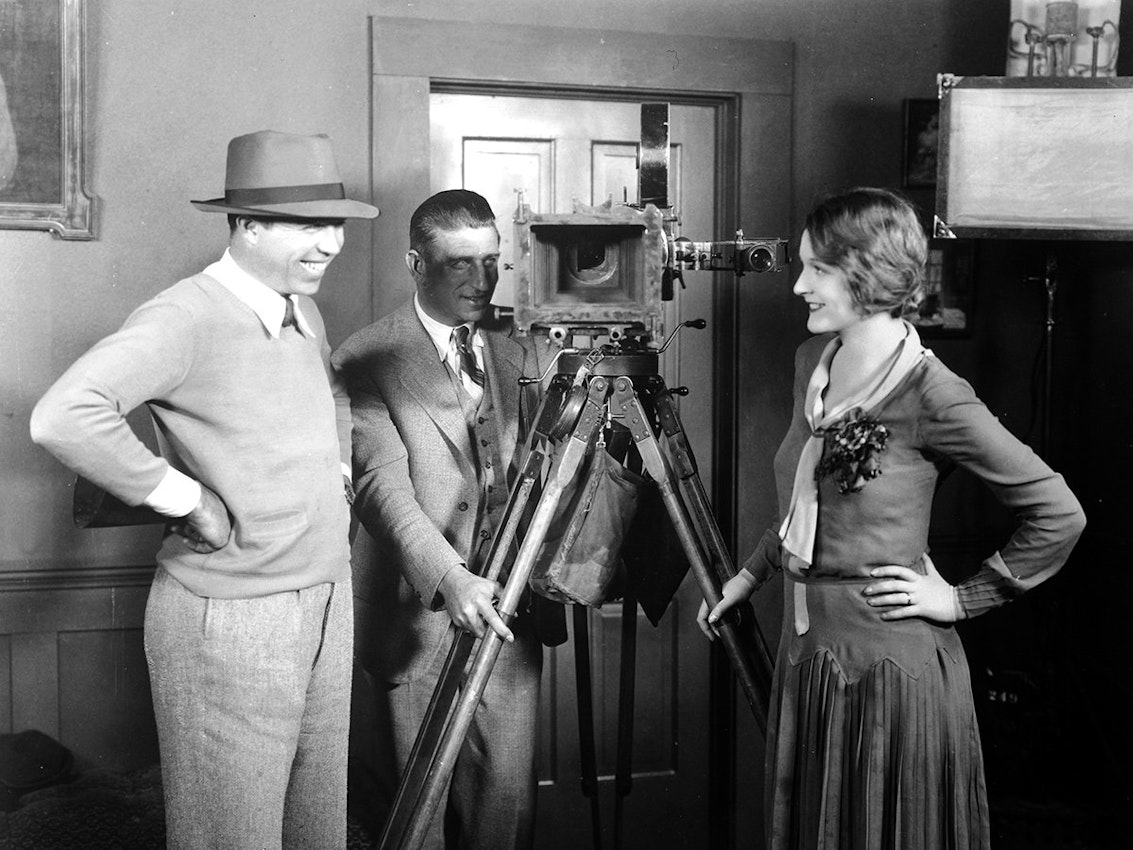 A man in a hat smiles while standing next to a camera on a tripod, with another man adjusting the camera and a woman smiling nearby.