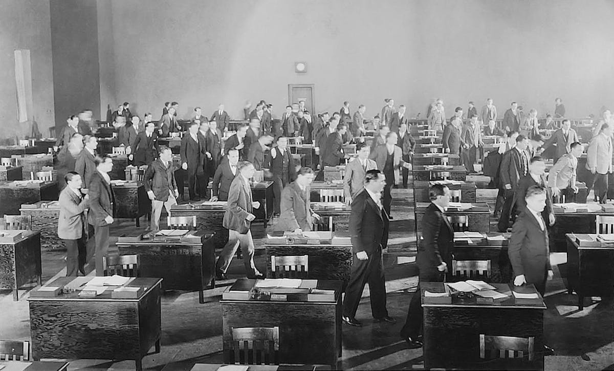 A large group of men in suits walk between rows of desks in an office space with high ceilings and minimal decoration.