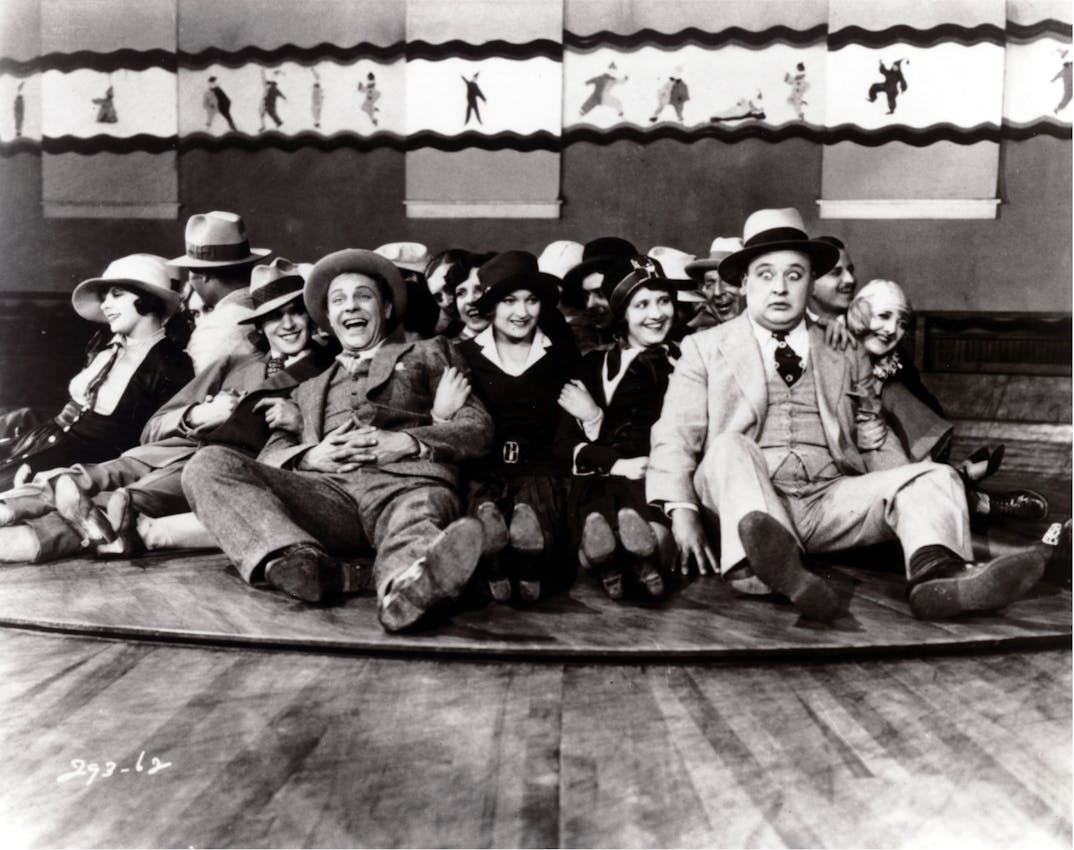 Several people, men and women in hats and formal clothing, are seated tightly together on a circular amusement ride. They appear to be enjoying themselves, smiling and laughing, while one man looks surprised or alarmed. Behind them, a mural with figures in motion decorates the wall.