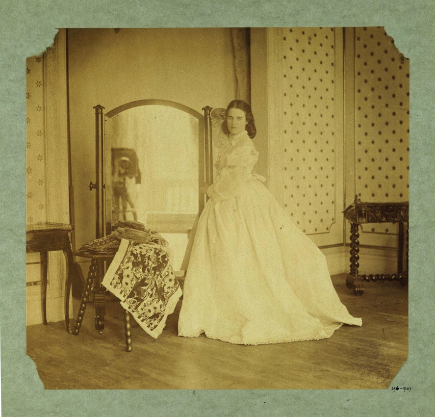 Sepia-toned photograph depicting a woman in a mid-19th century dress standing beside a mirror, with a patterned shawl draped over a small wooden table, and other period furniture in an interior space